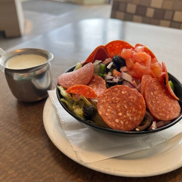 A salad with pepperoni, salami, tomatoes, olives, and lettuce, with a side of dressing.