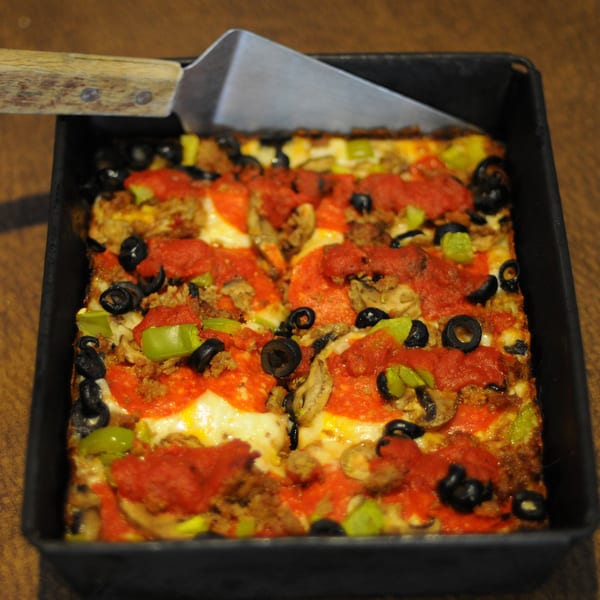 Deep dish pizza with pepperoni, sausage, olives, green peppers, and mushrooms in a square pan.