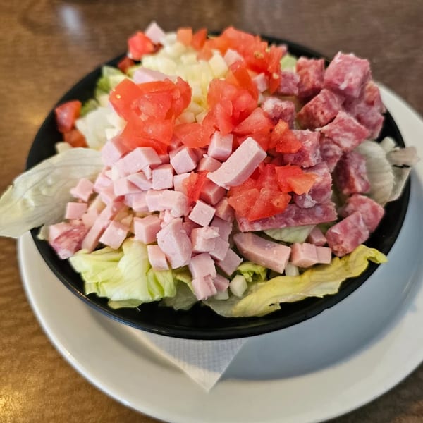 A black bowl of salad with lettuce, diced tomatoes, ham, and salami on a white plate.
