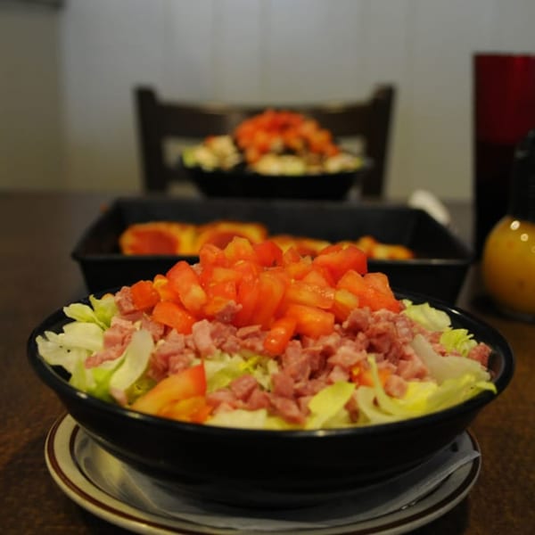 A close-up of a salad with lettuce, diced tomatoes, and ham, with a pizza in the background.